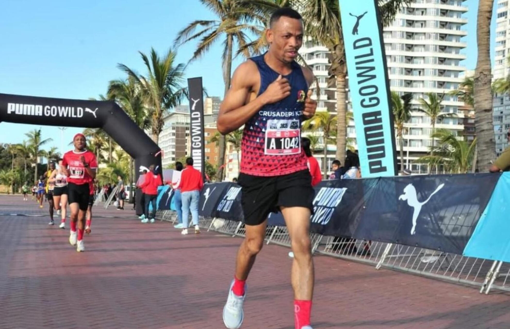 Eric Crossing the Finish Line Eric from Crusaders Running Club finishing strong at the Puma Go Wild race in Durban, wearing the club’s red and blue vest with bib A1042.